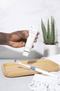 Hand holding a tube of toothpaste above a toothbrush on a wooden tray